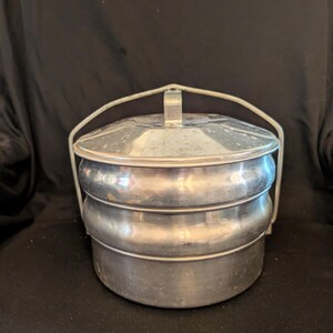 May include: A vintage, three-tiered metal lunch carrier with a handle. The carrier is made of a shiny, silver-colored metal and has a rounded shape. The handle is arched and attached to the top tier. The lunch carrier is likely used for carrying food.