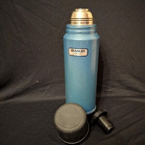 May include: A blue Stanley Sport thermos with a silver cap and a black stopper. The thermos has a textured finish and a Stanley Sport label. The black cap and stopper are placed next to the thermos.