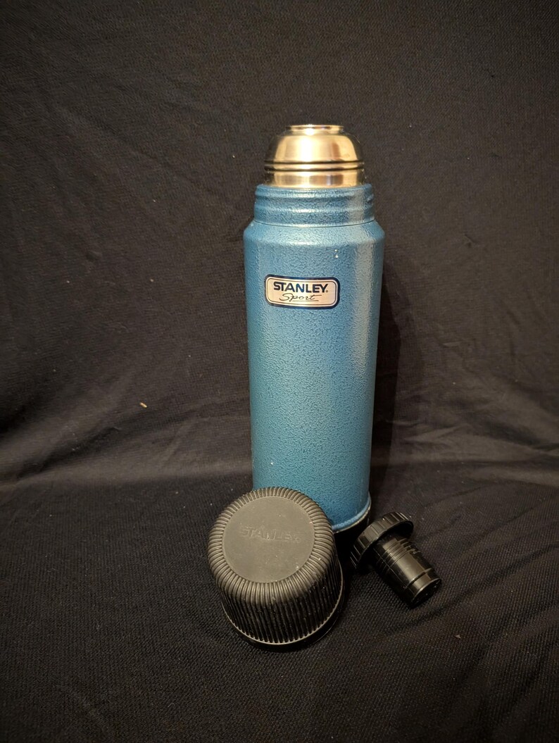 May include: A blue Stanley Sport thermos with a silver cap and a black stopper. The thermos has a textured finish and a Stanley Sport label. The black cap and stopper are placed next to the thermos.
