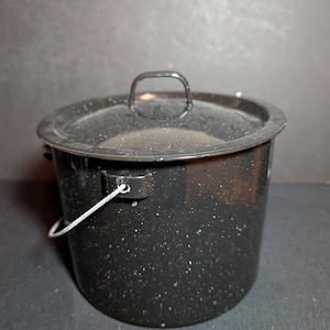 May include: A black enamel pot with a lid and a wire handle. The pot has a speckled pattern and a metal handle on the lid. The pot is designed for cooking and camping.