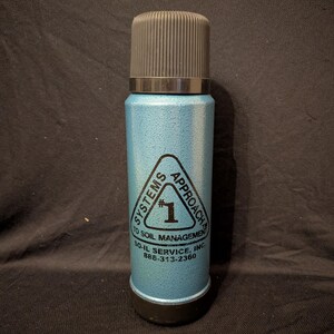 May include: A vintage, light blue thermos with a black cap and base. The thermos has a textured surface and features a triangular logo with the text "SYSTEMS APPROACH TO SOIL MANAGEMENT" and the number "1". The phone number 888-313-2360 is also visible.