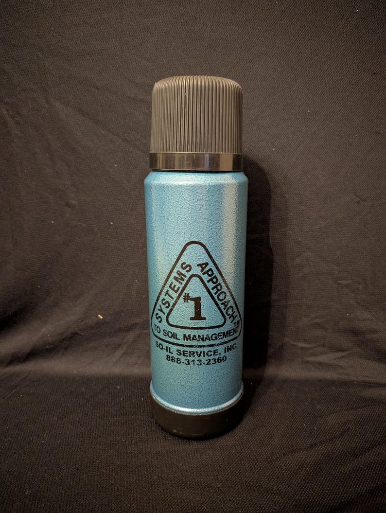 May include: A vintage, light blue thermos with a black cap and base. The thermos has a textured surface and features a triangular logo with the text "SYSTEMS APPROACH TO SOIL MANAGEMENT" and the number "1". The phone number 888-313-2360 is also visible.