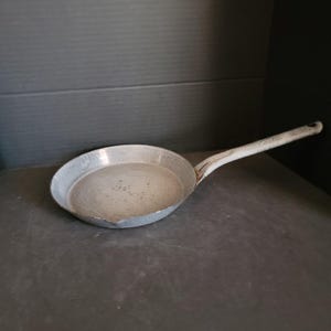May include: A small, well-worn, gray metal frying pan with a long, white handle. The pan has a rounded shape and a slightly textured surface, suggesting it has been used. The handle is attached to the pan at an angle.
