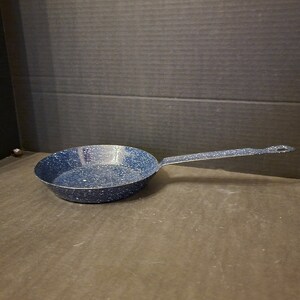 May include: A blue speckled enamel frying pan with a long handle. The pan is round with a slightly sloped side. The handle is attached to the pan with a riveted design. The pan is made of metal and has a speckled pattern.