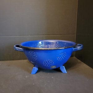 May include: A vibrant blue colander with small, evenly spaced holes. The colander has two side handles and stands on three small legs. The rim is black, and the overall design is simple and functional.