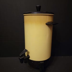 May include: Vintage coffee percolator with a two-tone design. The body is a light yellow with a darker yellow gradient, and the lid, handle, and base are black. Features a spigot and a black handle. A classic kitchen appliance.