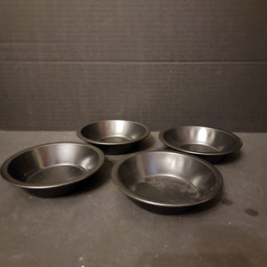 May include: Four small, round, black metal pie pans are arranged on a dark surface. The pans have a slightly sloped edge and a matte finish. They are empty and ready for use.