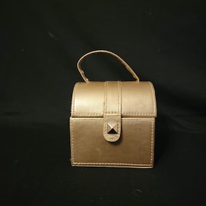 May include: A small, gold-colored box with a handle, likely a jewelry or trinket box. The box has a rectangular shape with rounded edges and a clasp with a decorative stud. The handle is small and arched.