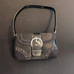 May include: A black handbag featuring a single shoulder strap, a central flap closure with a silver-toned buckle adorned with clear crystals, and decorative silver-toned metal grommets along the lower front corners with white contrast stitching.