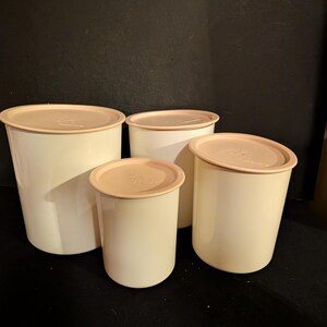 May include: Four off-white plastic food storage containers with peach-colored lids. The containers are cylindrical and vary in size, with the largest on the left and the smallest in the center. The lids have a slightly raised center.
