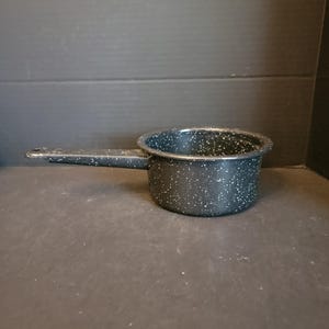 May include: A small, black speckled saucepan with a long handle. The pot is made of metal and has a rounded shape. The interior is a solid black color. The handle extends to the left, and the pot is sitting on a dark surface.