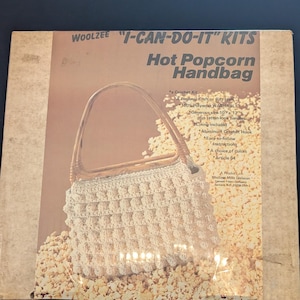 May include: A vintage "I-CAN-DO-IT" kit by Woolzee featuring a crochet pattern for a "Hot Popcorn Handbag." The kit includes instructions, an aluminum crochet hook, and a choice of colors. The handbag is shown in a cream color with rattan-look handles.