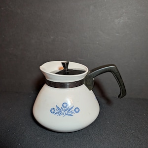 May include: White coffee pot with a black handle and lid. The pot has a floral design in blue. The pot is made of enamel and has a black band around the middle. The handle is curved and attached to the side of the pot.