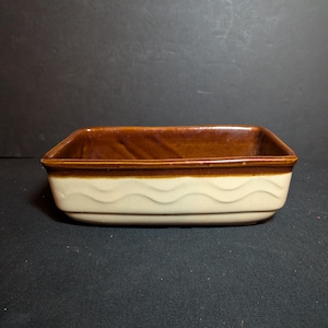 May include: A rectangular ceramic baking dish featuring a two-tone design. The interior and the upper rim are finished in a glossy dark brown glaze, while the exterior sides are a cream color with a horizontal embossed wavy line pattern.