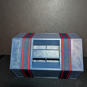 May include: A blue plastic Rolykit storage organizer featuring a rectangular shape with two vertical dark blue and red fabric straps. The central section displays the embossed text ROLYKIT and includes horizontal dividers for organized storage.