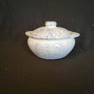 May include: A small, round ceramic bowl with a lid. The bowl and lid are white with a speckled blue pattern. The lid has a small knob on top for easy removal. The bowl has two small handles on either side.
