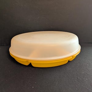 May include: A vintage Tupperware container with a yellow base and a translucent white lid. The container has a rounded, oval shape and is designed for food storage. The lid fits snugly onto the base, creating an airtight seal.
