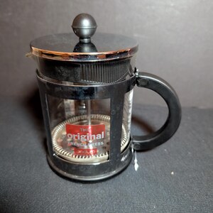 May include: A French press coffee maker with a black frame, handle, and lid. The clear glass carafe has a stainless steel filter and a red label that reads "THE ORIGINAL FRENCH PRESS". The coffee maker is designed for brewing coffee.