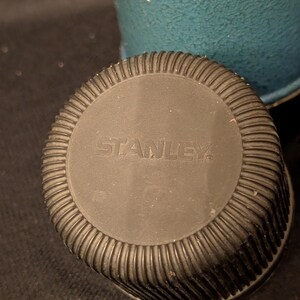 May include: Close-up of a black Stanley thermos lid with a ribbed edge. The word "STANLEY" is embossed in the center. A teal-colored thermos body is visible in the background.