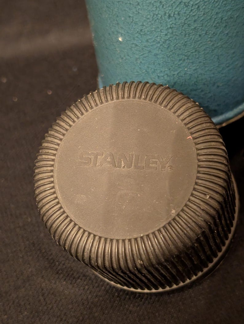 May include: Close-up of a black Stanley thermos lid with a ribbed edge. The word "STANLEY" is embossed in the center. A teal-colored thermos body is visible in the background.