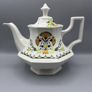 May include: White ceramic teapot with a geometric design. The teapot features a lid with a finial, a spout, and a square handle. The body is decorated with orange, yellow, and green floral patterns. The teapot has a hexagonal base.