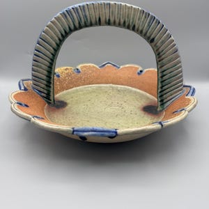 May include: A ceramic basket with a scalloped edge and a handle. The basket features a light green interior, an orange and brown rim, and a cream-colored exterior with blue accents. The handle is textured with blue and green stripes.