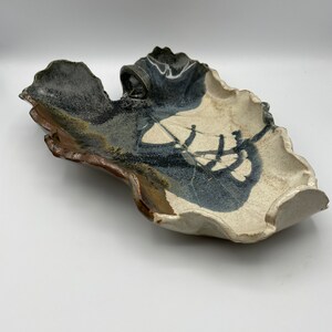 May include: A ceramic serving dish with an irregular, organic shape. The dish is glazed in shades of white, brown, and black, with a textured surface.