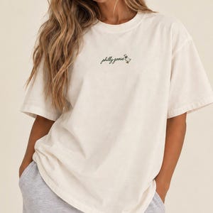 May include: An off-white, short-sleeved t-shirt featuring a crew neckline and a relaxed fit. The center chest displays small, dark green cursive text that reads "philly goose" accompanied by a tiny, detailed illustration of a goose wearing a bow.
