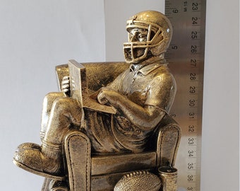 May include: A gold-colored trophy of a football player sitting in a chair with a football and a drink in his hands. The trophy is on a gold-colored base.
