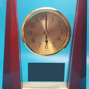 May include: A gold-coloured clock with black hands and black Roman numerals on a white face. The clock is set to 12:00 and is framed in a clear glass case with a dark red wooden base.