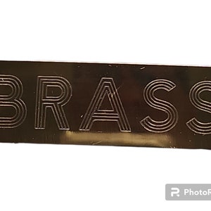 May include: A black metal sign with the word "BRASS" engraved in a stylised font.