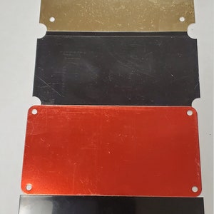 Puede incluir: Una placa negra con el texto "CUSTOM ENGRAVED PLAQUE 2"x4" 5 LINES 30 CHARACTERS PER LINE". La placa se muestra con otras tres placas en oro, negro y rojo.
