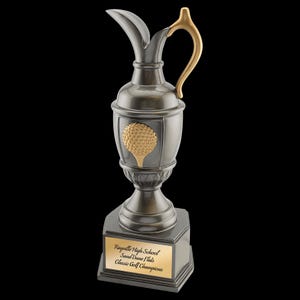 Może przedstawiać: Puchar golfowy w kolorze cyny ze złotym uchwytem i emblematem piłki golfowej. Puchar ma kształt dzbanka i stoi na kwadratowej podstawie ze złotą tabliczką z napisem "Rayville High School Sand Dune Flats Classic Golf Champions."