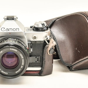 May include: A silver Canon AE-1 Program camera with a black lens and a brown leather case. The camera has the Canon logo and lens information visible. The case is brown with the Canon logo in gold.