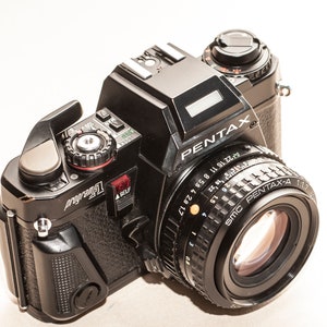 Könnte beinhalten: Eine schwarze Pentax-Kamera mit einem Objektiv mit der Aufschrift SMC PENTAX-A 1:1.7. Die Kamera hat einen silbernen Objektivring mit Zahlen und ein schwarzes Gehäuse. Das Wort "PENTAX" ist auf der Vorderseite der Kamera aufgedruckt.