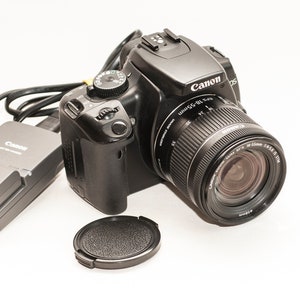 May include: A black Canon EOS digital SLR camera with a lens, lens cap, charger, and power adapter. The lens is a Canon EF-S 18-55mm f/3.5-5.6 IS STM. The camera and accessories are on a white background.