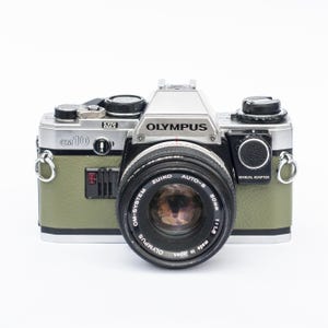 Puede incluir: Una cámara de película de 35mm Olympus OM-10 con una parte superior de metal plateado y un cuerpo texturizado de color verde oliva. La cámara cuenta con un objetivo negro Zuiko Auto-S 50mm 1:1.8, un adaptador manual y enganches plateados.