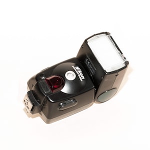 Peut inclure: Un flash Nikon Speedlight SB-80DX noir avec une tête de flash blanche et un voyant LED rouge. Le flash a un corps en plastique noir et un pied en métal argenté.