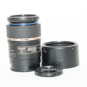 May include: A black Tamron SP Di 90mm macro lens with a lens hood and lens cap. The lens has gold lettering and a blue accent ring. The lens cap and hood are black. The lens has a focus distance scale.