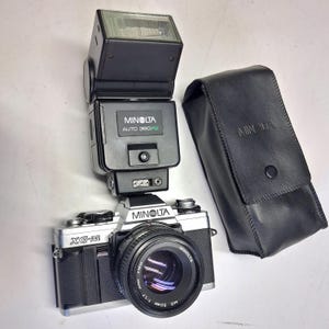 Puede incluir: Una cámara de película Minolta XG-M de 35 mm con un objetivo de 50 mm, un flash negro y una funda de cuero negra. La cámara y el flash son negros y plateados. El objetivo tiene una apertura de 1:1,7.