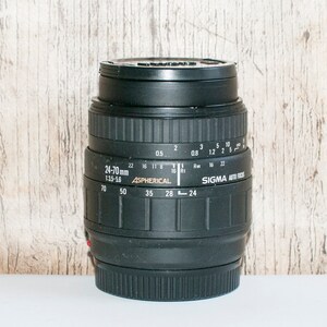 May include: A black Sigma 24-70mm f/3.5-5.6 Aspherical lens with a silver zoom ring and aperture markings. The lens is designed for 35mm film cameras.