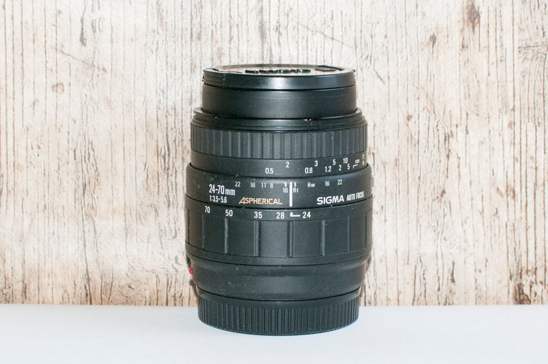 May include: A black Sigma 24-70mm f/3.5-5.6 Aspherical lens with a silver zoom ring and aperture markings. The lens is designed for 35mm film cameras.