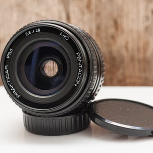 May include: A black Prakticar PM 2.8/28 MC camera lens with a lens cap. The lens has the word "Pentacon" printed on it. The lens is sitting on a white surface with a wooden background.