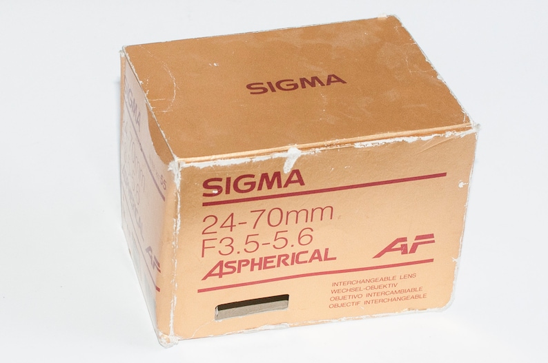 May include: A gold box with the text "SIGMA 24-70mm F3.5-5.6 ASPHERICAL AF" printed on it. The box is for an interchangeable lens.