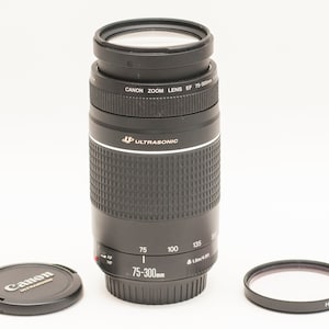 May include: A black Canon Zoom lens EF 75-300mm with a lens cap and a filter. The lens has a textured grip and markings for 75, 100, 135, and 200mm. The lens cap has the Canon logo.
