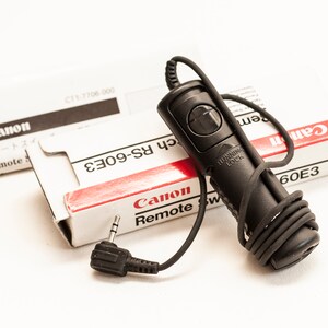 May include: A black Canon RS-60E3 remote switch with a black cord and a black plug. The remote switch has a button with the words "Running Lock" printed on it. The remote switch is packaged in a white box with red and black text that reads "Canon Remote Switch RS-60E3".