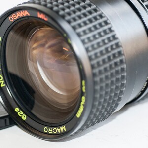 Può includere: Un obiettivo per fotocamera nero con una fascia argentata e il testo "MACRO" e "628" stampato in giallo. L'obiettivo ha anche il testo "OSAW A" e "NO. 1025090" stampato in giallo. L'obiettivo è stato prodotto in Giappone.