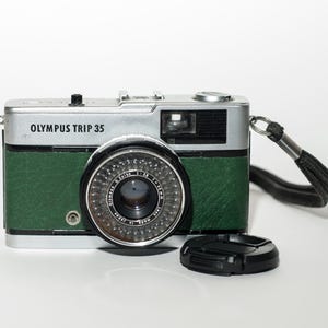 Op de afbeelding: Een Olympus Trip 35 camera met een zilveren bovenkant, groene kunstleren behuizing en een zwarte lens. De camera heeft een zwarte lensdop en een zwarte polsriem. De tekst "OLYMPUS TRIP 35" staat op de voorkant.