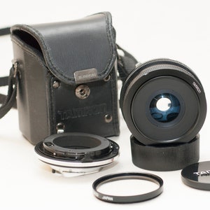 Puede incluir: Un objetivo de cámara Tamron negro con una tapa de objetivo, filtro y anillo adaptador a juego. También se incluye una funda de cuero negro con correa para el hombro. El objetivo está sobre un fondo blanco.