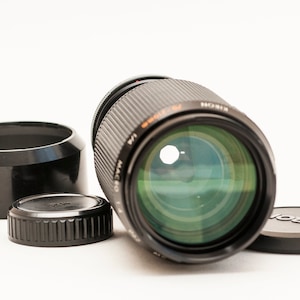 Puede incluir: Un objetivo de cámara negro con un elemento de vidrio teñido de verde, acompañado de un parasol y una tapa de objetivo. El objetivo tiene texto naranja que dice "MACRO 1:4". La tapa del objetivo tiene la marca "MINOLTA".